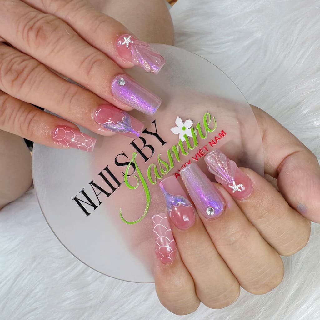 Bộ móng nàng tiên cá với hiệu ứng ánh nhũ hồng tím, họa tiết vỏ sò – vây cá và đính đá ngọc trai, mang phong cách fantasy dịu dàng, do học viên Jasmine thực hiện